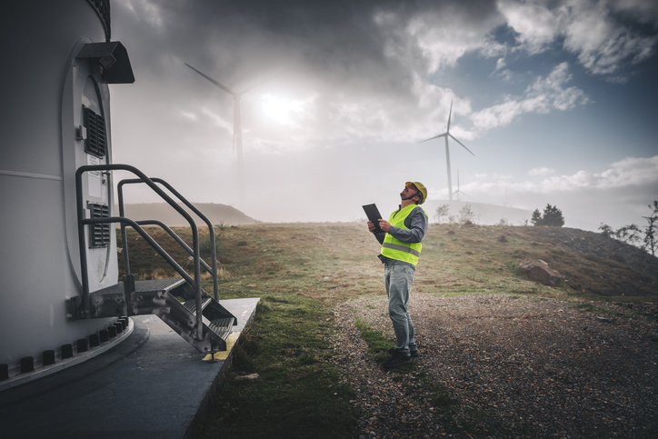 Portrait of young caucasian technician engineer man wearing yellow worker headwear standing, working, checking wind farm field system and looking up verify at wind turbine while using a radio station energy plan by wind turbine power generation station